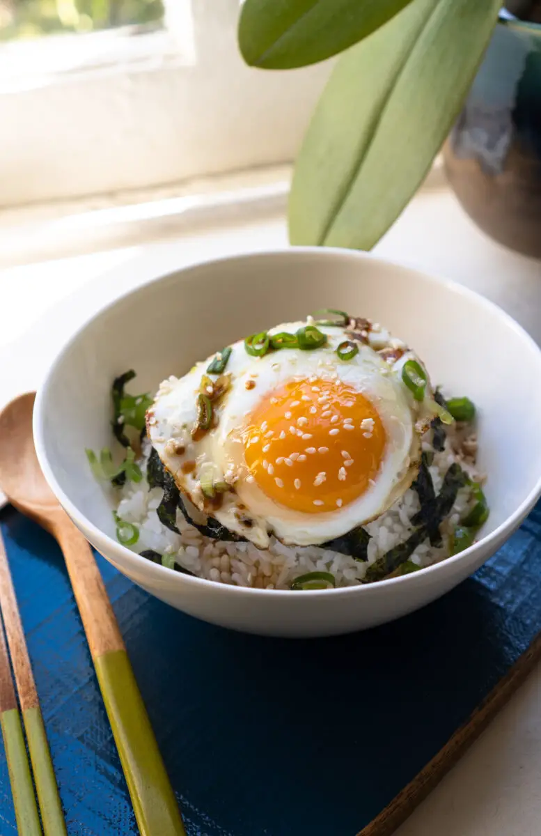 A side shot of gyeran bap. This rice bowl contains seaweed strips, green onions, and a sunny side up egg. The white bowl sits on a blue tray. 