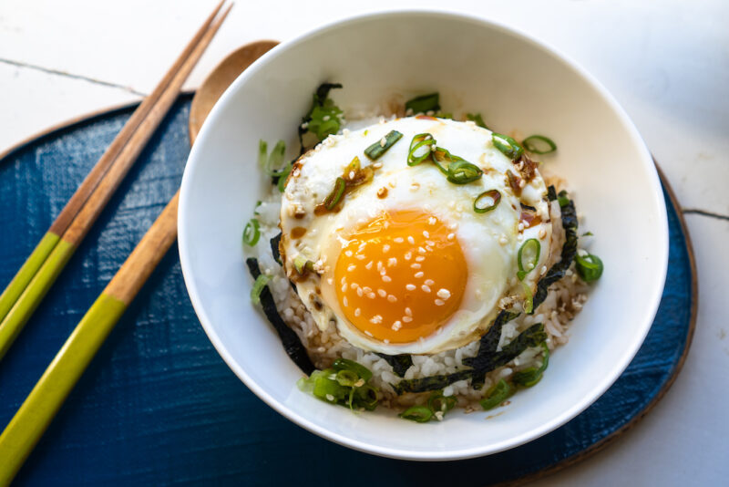 An overhead shot of gyeran bap. Thia bowl of rice has a sunny side egg sitting on top of rice. Green onions and seaweed strips are in the bowl. The bowl sits on a blue tray next to chop sticks. 