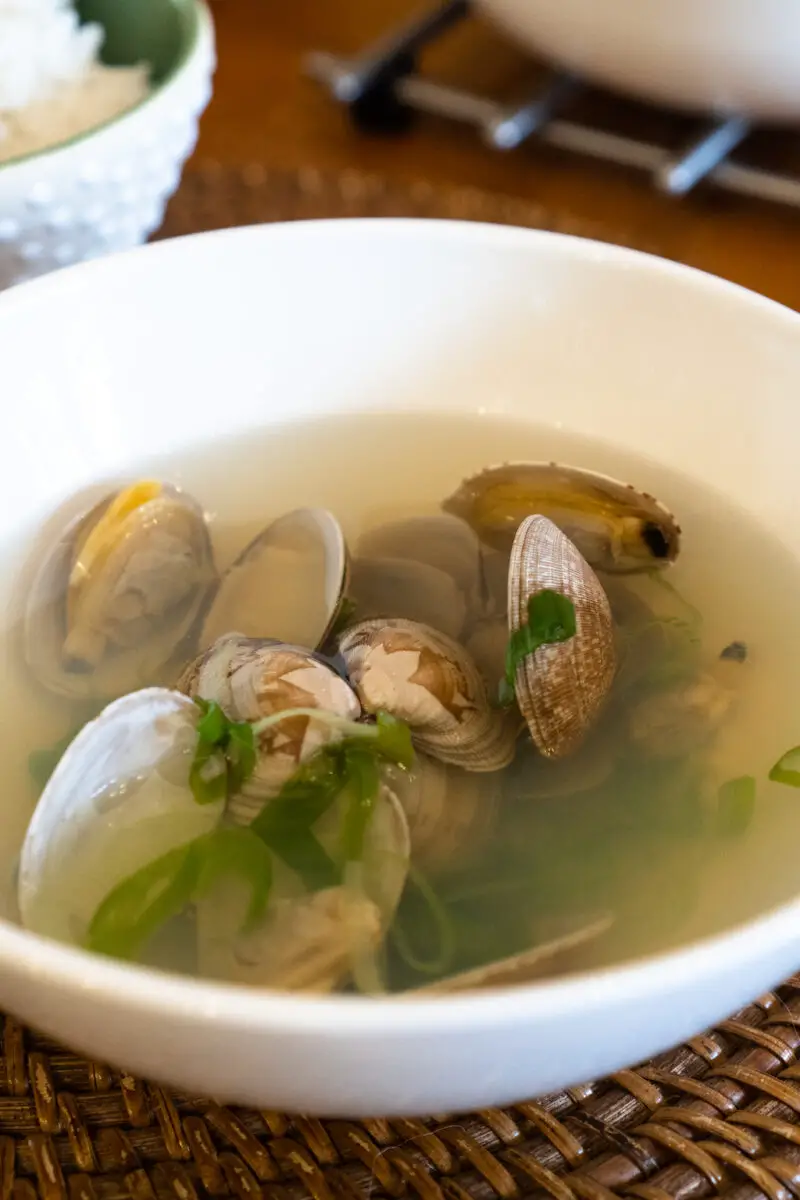 A closeup side shot of clams in a clear broth. In the soup there are also thinly sliced green onions and Korean peppers.