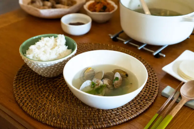 A bowl of Korean clam soup sit nexts to a bowl of white rice on a table mat. In the background sits the pot of soup next to kimchi, ssamjang, radish, and more.