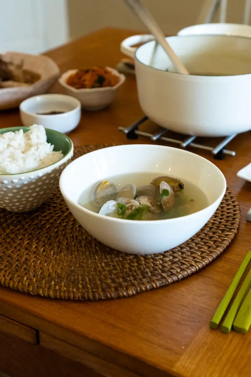 A side shot of a bowl of Korean clam soup next to a bowl of white rice. The rice and soup sits on a table mat. A full Korean meal sits in the background.
