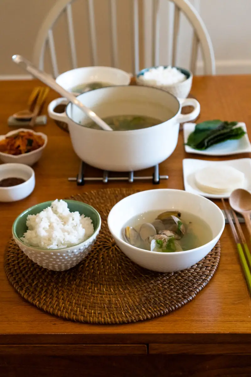 A full Korean meal on a table. The meal includes jogaetang, white rice, pickled radish, perilla leaves, and kimchi. 