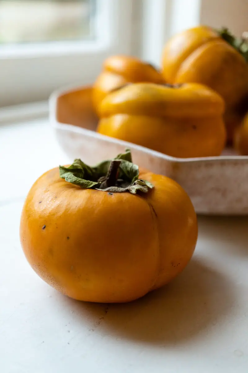 A persimmon sits in the middle of the photo on a white window sill. A bowl of the same fruit sit in the background.