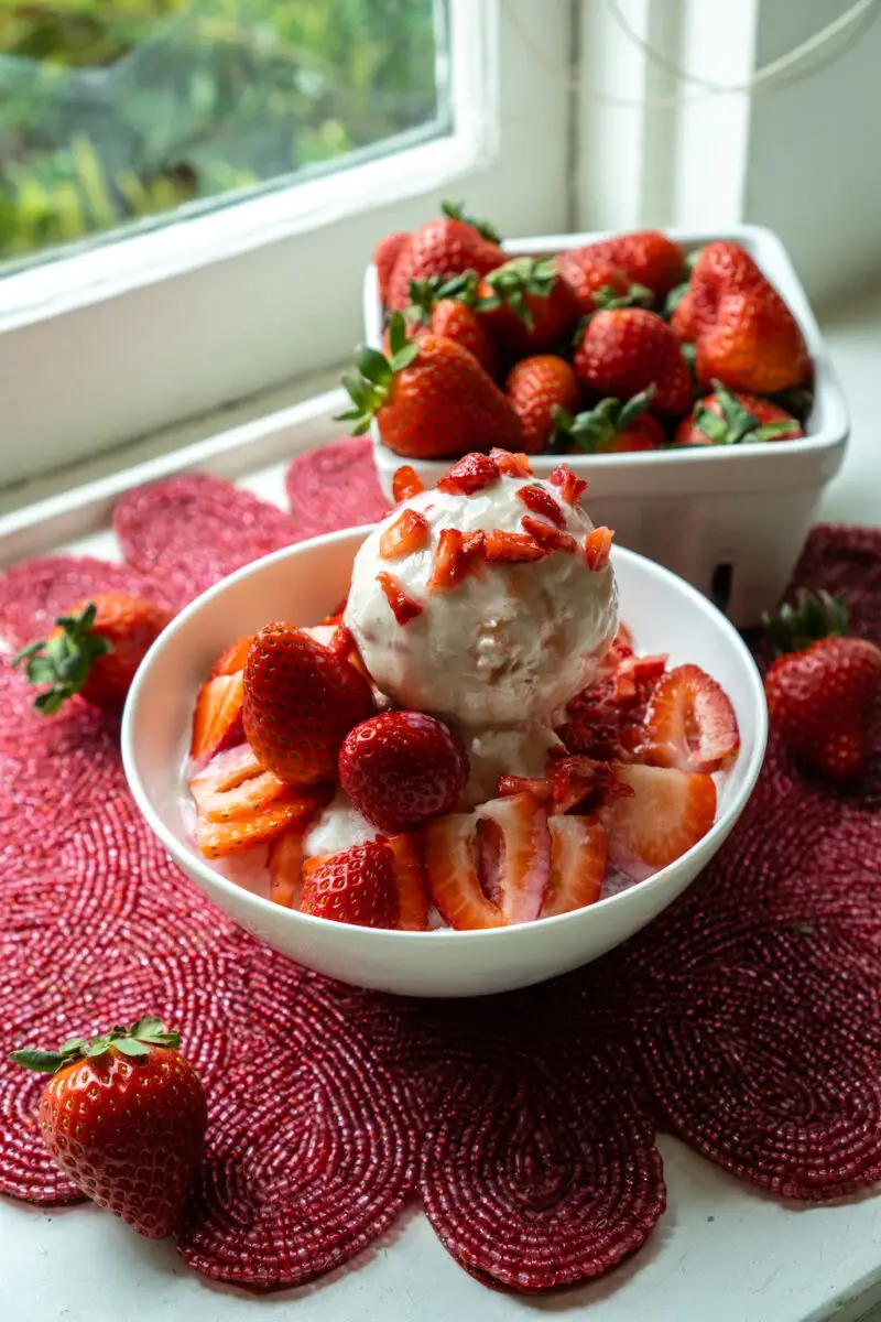 Another shot of Korean strawberry bingsu topped with strawberry ice cream in a white bowl. It sits on a red place mat. A container of strawberries sit on the background.
