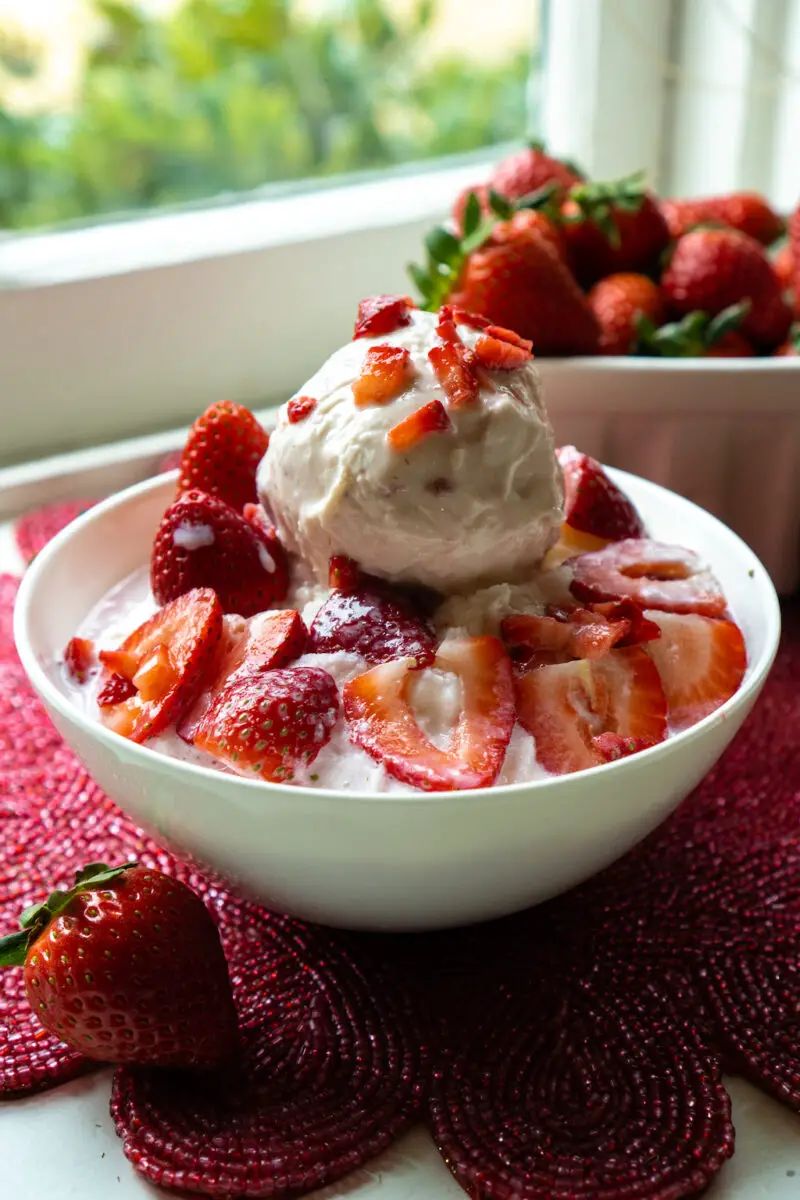 Korean strawberry shaved ice topped with strawberry ice cream in a white bowl. It sits on a red place mat. A container of strawberries sit on the background.