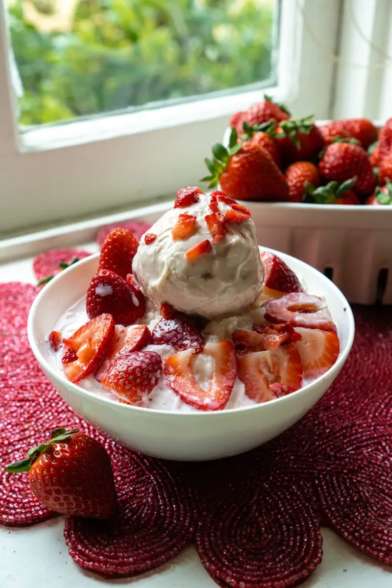 A side shot of Korean strawberry bingsu topped with strawberries. The bingsu sits on a red placemat next to a bowl of strawberries.