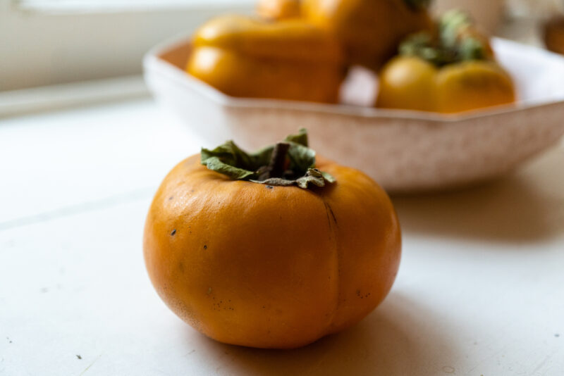 A side shot of a Korean persimmon fruit sitting on a window sill. In the background sits a bowl of more persimmons. 