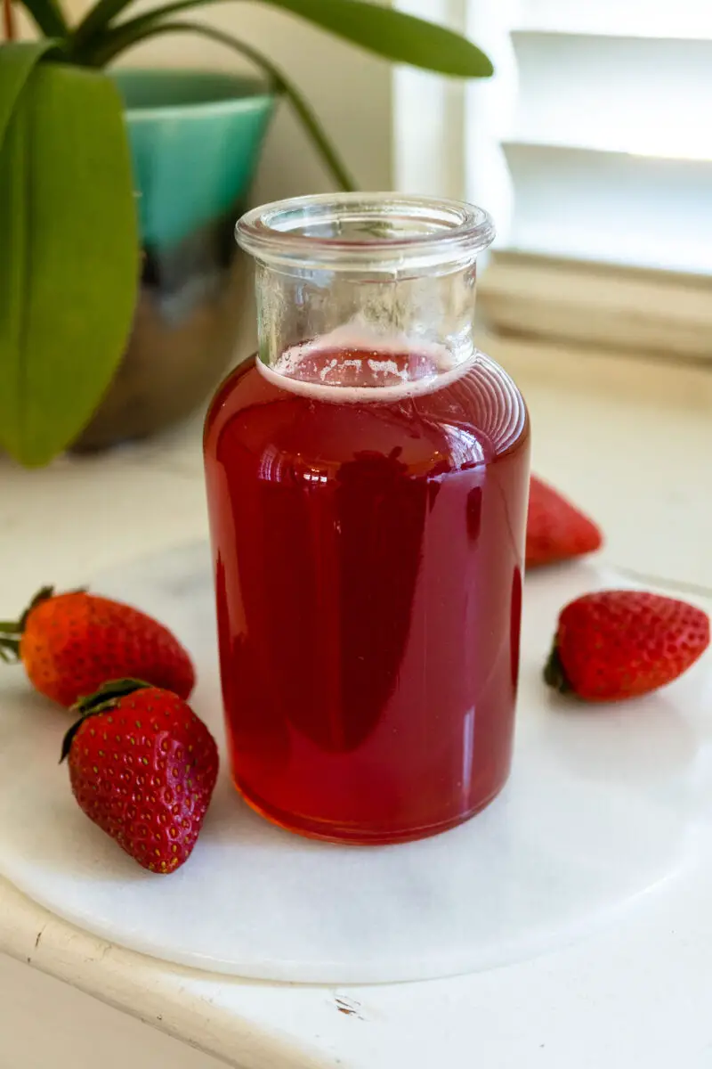 A closeup side shot of strawberry simple syrup in a glass container next to fresh strawberries. 