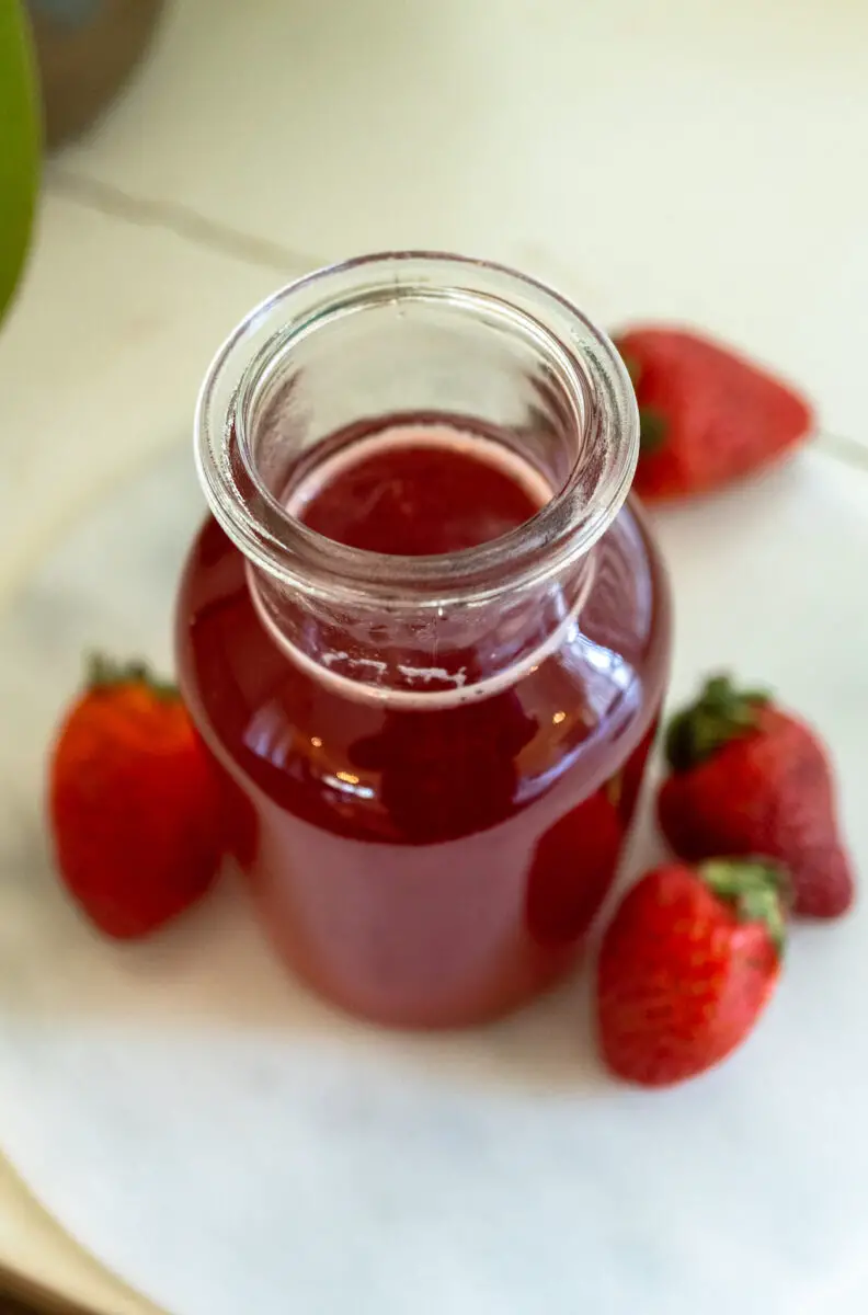 Bright red syrup made from fresh strawberry fruit. Strawberries sit around the glass container. 