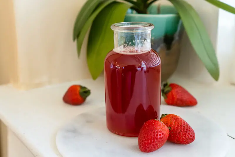 Bright red liquid in a glass jar made from fresh fruit. The mixture sits on a white window sill on a circle marble serving platter. 