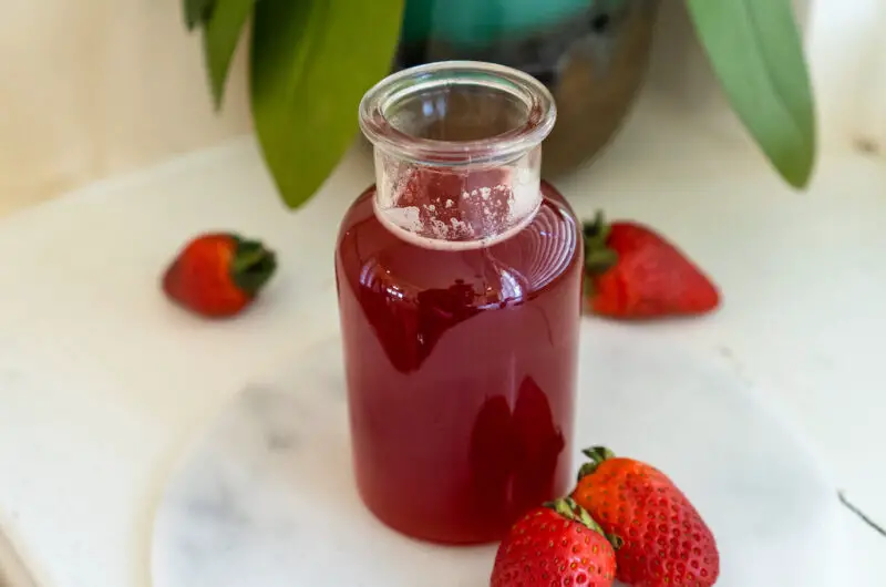 A closeup side shot of strawberry simple syrup in a glass container next to fresh strawberries. 