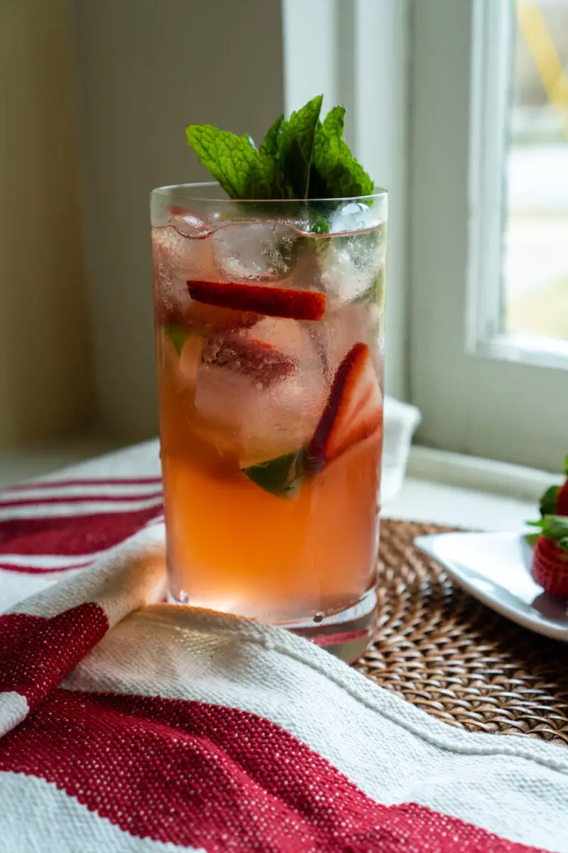 A strawberry soju cocktail sitting on a window sill. The cocktail sits on a mat and red and white linen. The strawberries sit on a plate next to a bottle of soju.