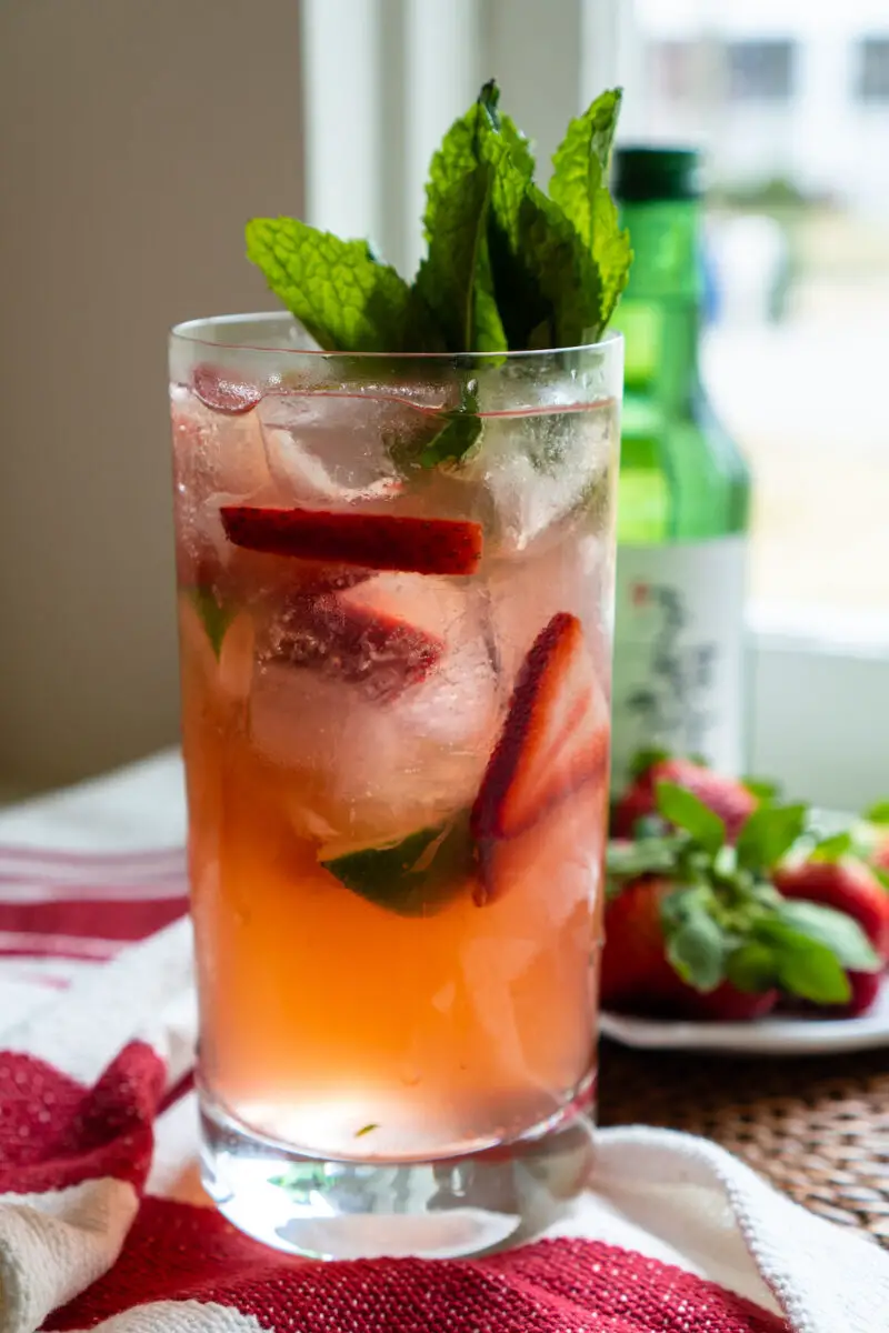 A photo of soju strawberry cocktail in a highball glass. Mint sticks out of the top of the glass. The glass is filled with sliced strawberries as well. A green bottle of soju sits in the background. 