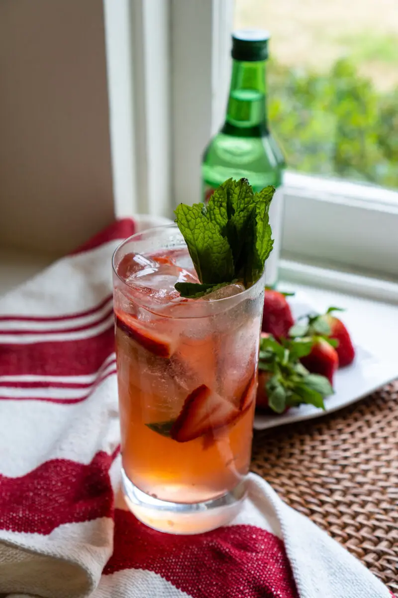 A strawberry soju cocktail sitting on a window sill. The cocktail sits on a mat and red and white linen. The strawberries sit on a plate next to a bottle of soju.