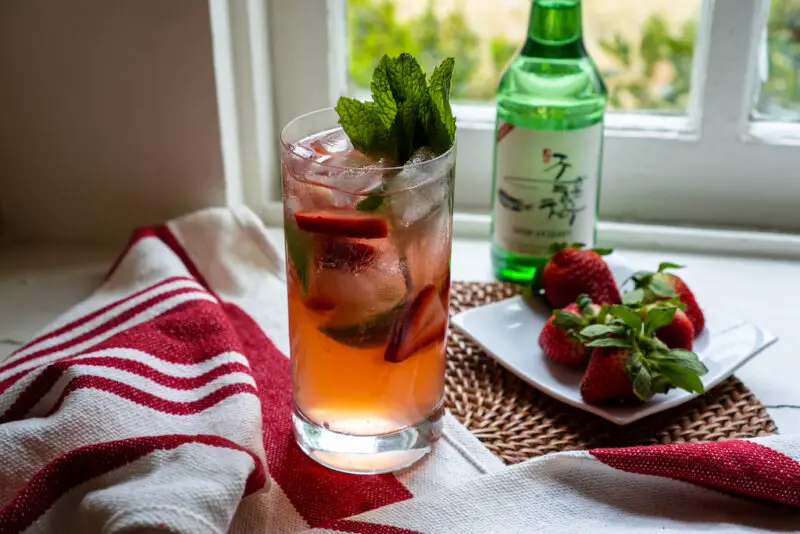 A strawberry soju cocktail sitting on a window sill. The cocktail sits on a mat and red and white linen. The strawberries sit on a plate next to a bottle of soju.