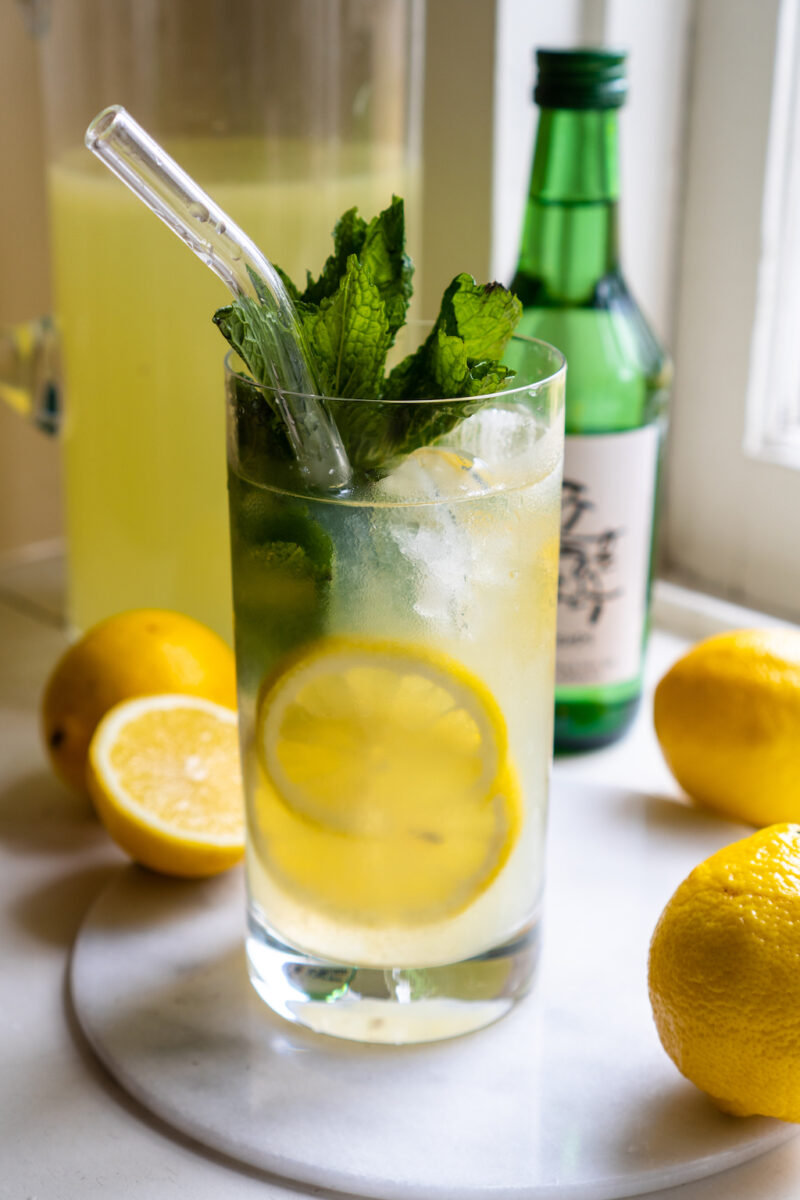 A side shot of soju lemonade cocktail. The drink sits next to lemons, lemonade, and a bottle of soju. Mint is in the drink. A straw sits in the highball drink. 