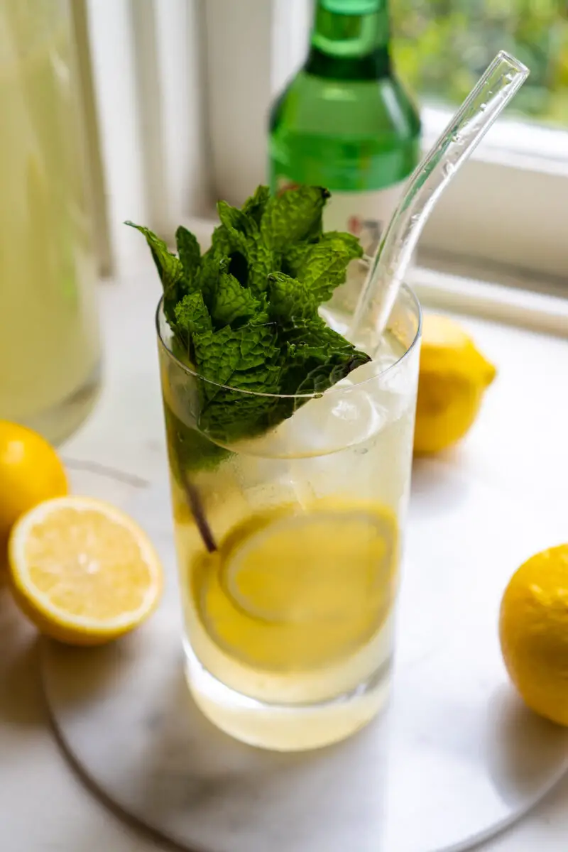 A side shot of this soju lemonade cocktail. The drink is in a high ball glass with a straw. It sits next to lemons, lemonade, and a bottle of soju. Mint is in the drink. 