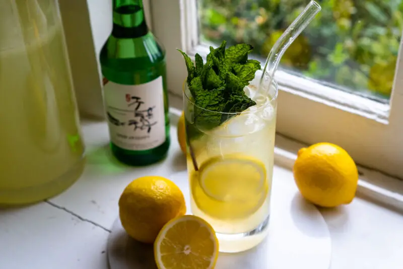 A side shot of soju lemonade cocktail on a window sill. The drink sits next to lemons, lemonade, and a bottle of soju. Mint is in the drink.