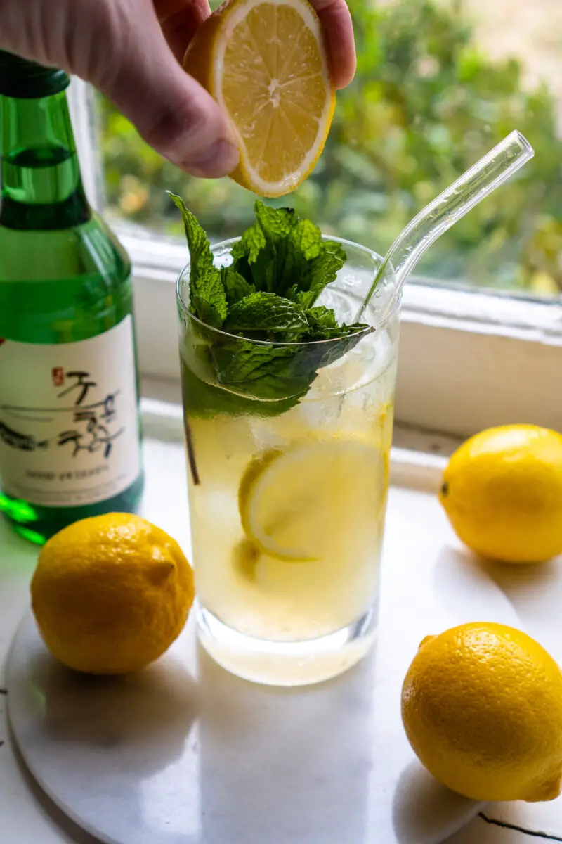 A side shot of me squeezing a lemon over my soju cocktail. The drink sits next to lemons, lemonade, and a bottle of soju. Mint is in the drink. 