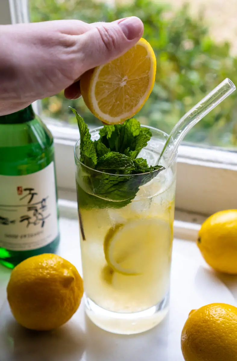 A side shot of me squeezing a lemon over my soju cocktail. The drink sits next to lemons, lemonade, and a bottle of soju. Mint is in the drink. 