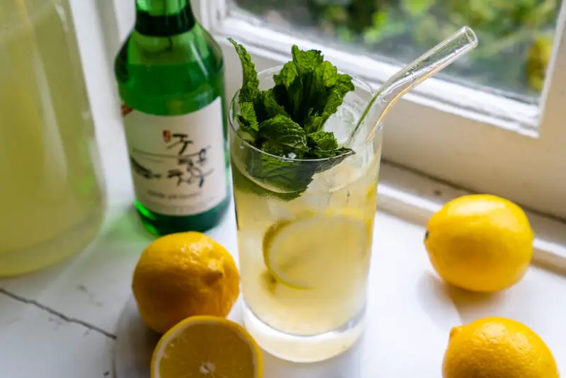 A side, overhead shot of soju lemonade cocktail on a window sill. The drink sits next to lemons, lemonade, and a bottle of soju. Mint is in the drink. 