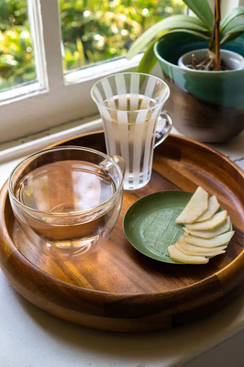 A side shot of two glasses of tea next to slices of pear on a wooden tray. 