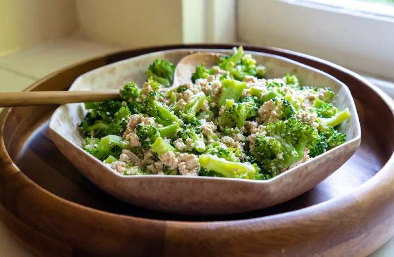 A photo of a Korean broccoli side dish  in an octagon bowl. The bowl sits in a wooden round tray. A wooden serving spoon its in the bowl.