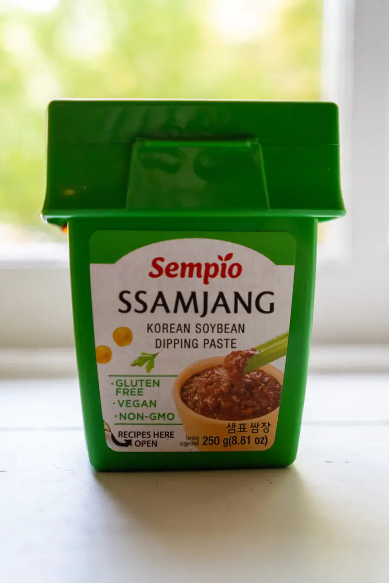 A closeup side shot of Sempio gluten-free ssamjang. This Korean ssamjang is in a bright green container on a window sill. 