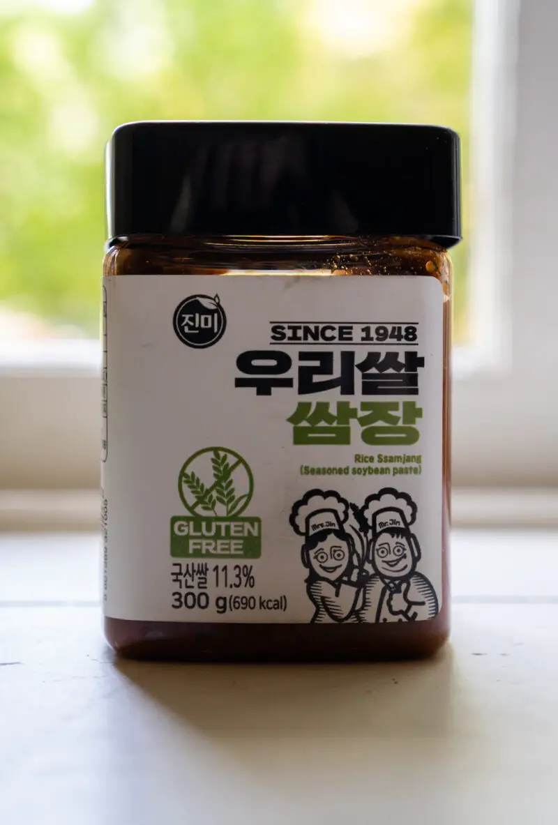 A closeup side shot of Jinmi gluten-free ssamjang. This Korean ssamjang is in a clear container on a window sill. You can see the dark brown sauce in the container.
