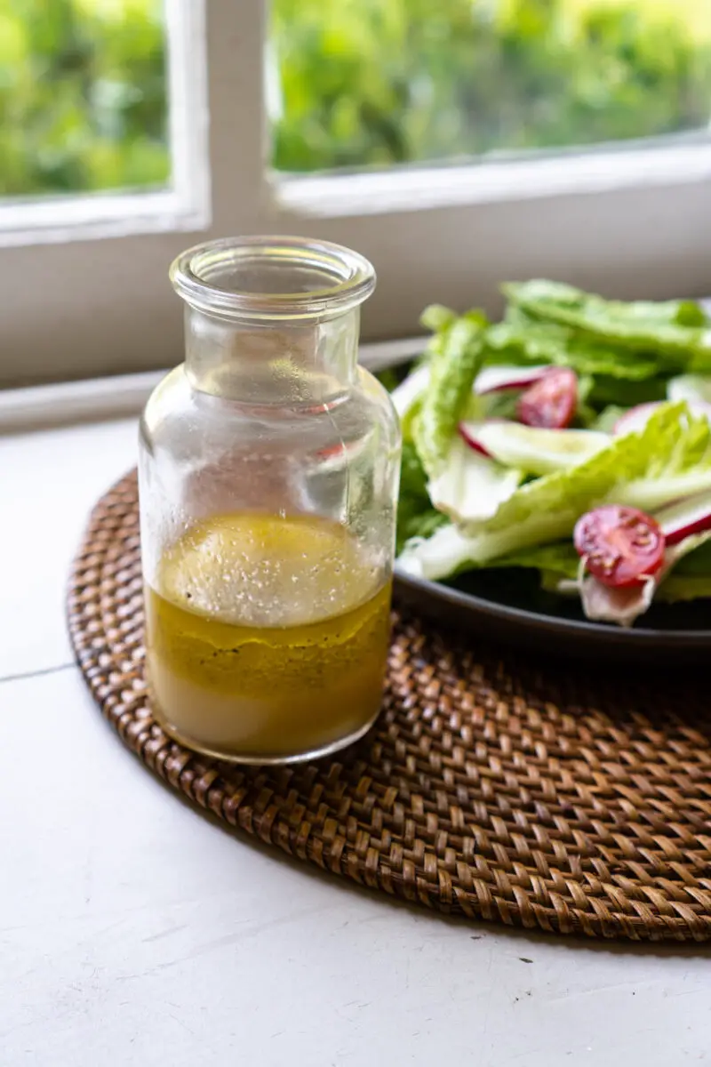 Korean yuzu vinaigrette dressing in a glass container. A salad with romaine lettuce, tomatoes, radish, and cucumber sit in the background. The salad and dressing sit on a table mat with a window in the background. 