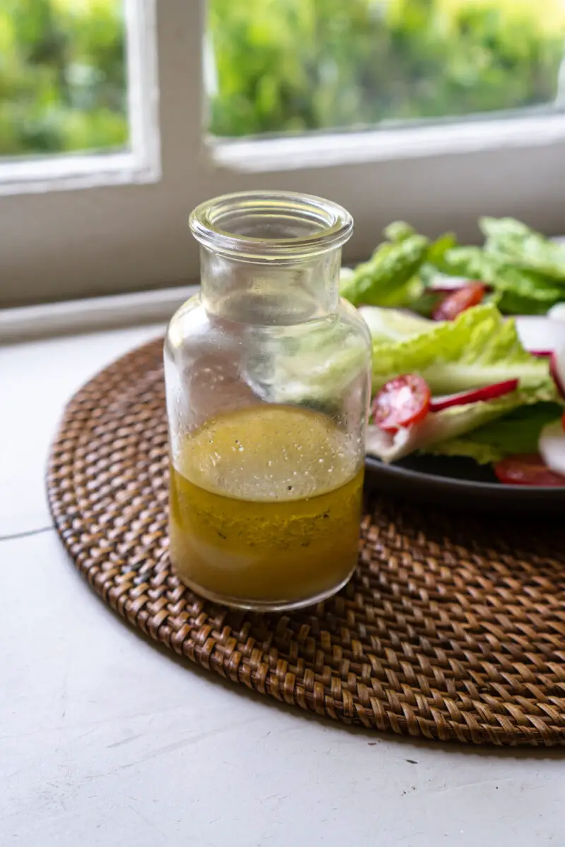 A side shot of Korean yuzu dressing in a glass container. A salad with romaine lettuce, tomatoes, radish, and cucumber sit in the background. The salad and dressing sit on a table mat with a window in the background.