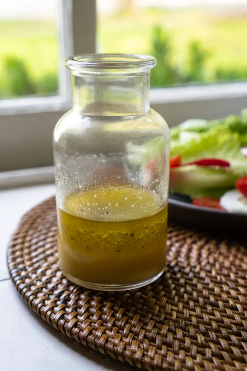 A closeup shot of Korean yuzu vinaigrette dressing in a glass container. A salad with sits in the background. The salad and dressing sit on a table mat. 