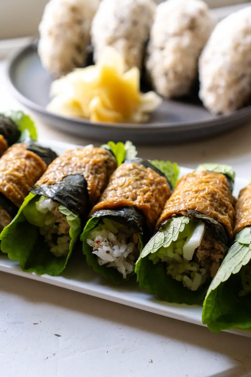 A closeup shot of rolled yubuchobap. The rolls are filled with rice, perilla leaves, and more! Rice balls and ginger sits in the background. 
