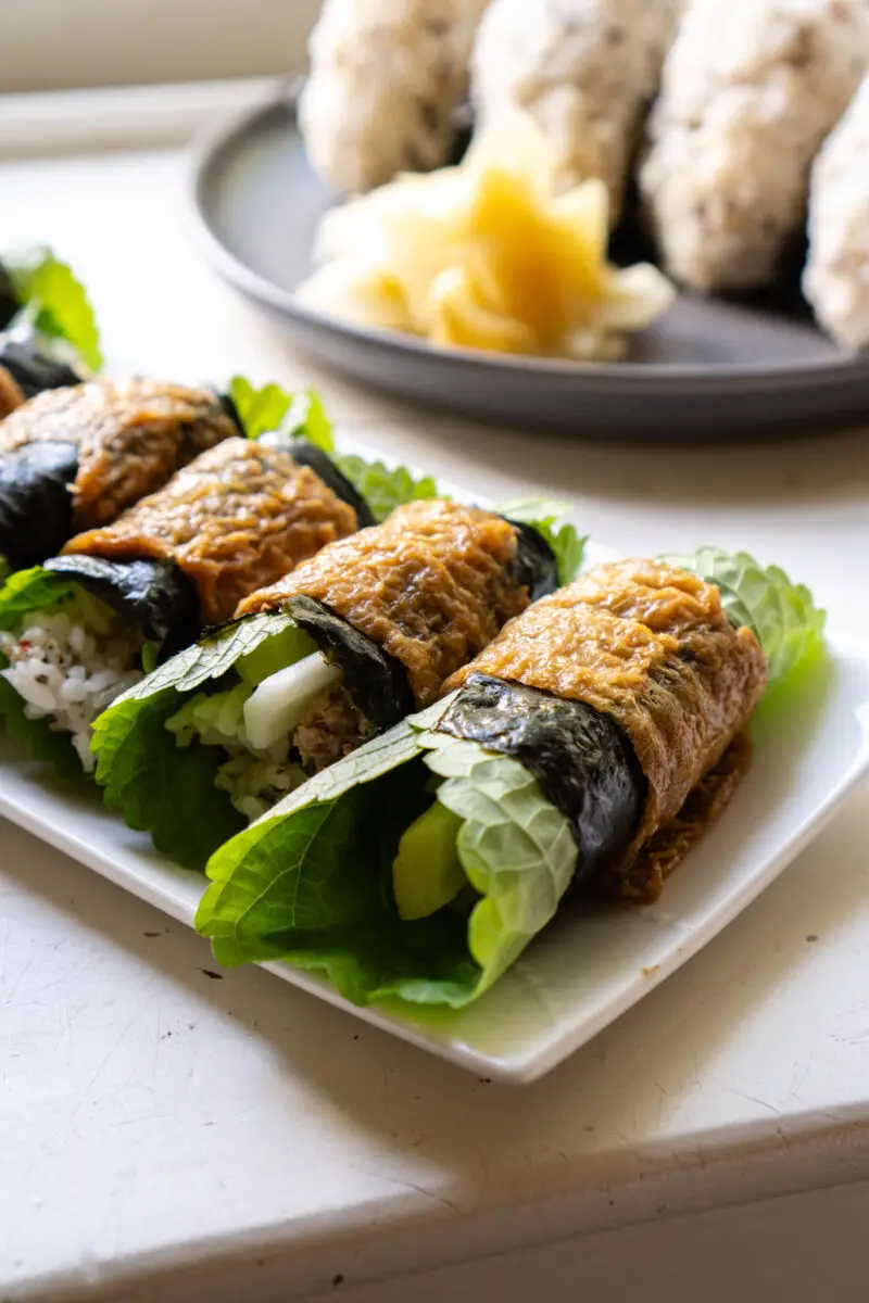 A closeup shot of rolled yubuchobap. The rolls are filled with rice, perilla leaves, and more! Rice balls and ginger sits in the background. 