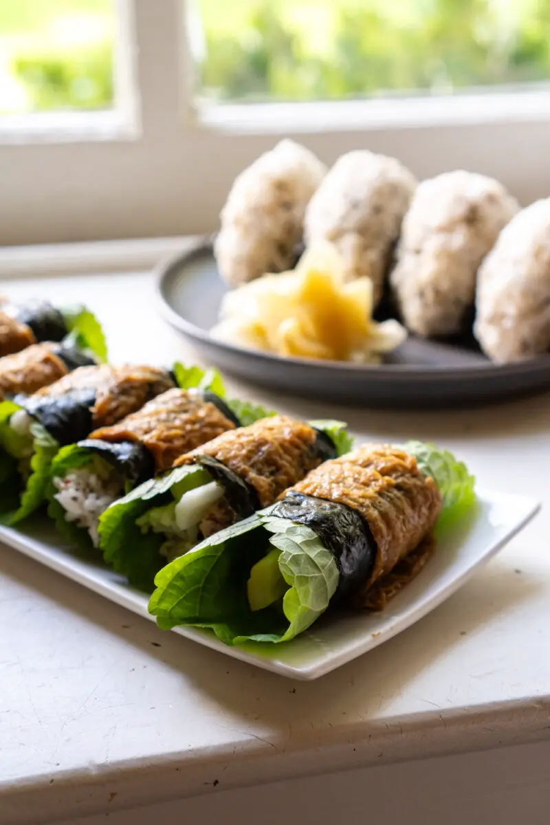 A closeup side shot of rolled Korean-style inari rolled sushi. The rolls are filled with rice, perilla leaves, and more! Rice balls and ginger sits in the background.