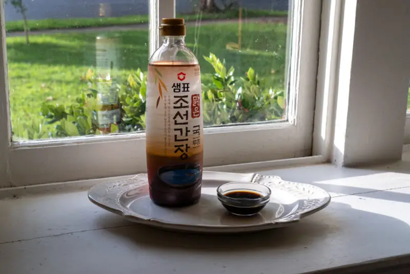 Korean guk ganjang in a clear bowl on a window sill. It sits next to the brand container on a cream tray. 