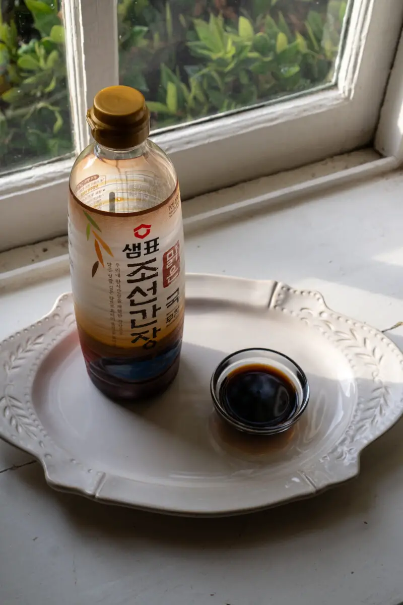 Korean soup soy sauce in a clear bowl on a window sill. It sits next to the brand container on a cream tray. 