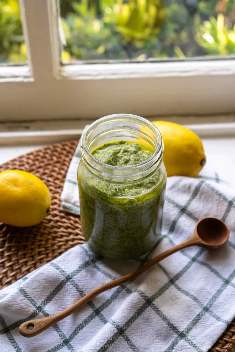 A side shot of bright green pesto in a glass jar. The jar sits next to lemons, a cloth napkin, and a wooden spoon. Everything sits on a white window sill.
