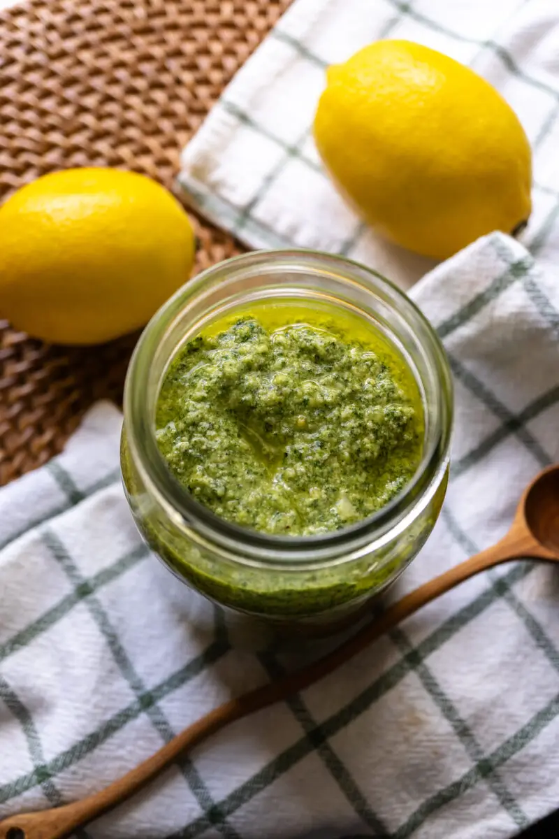 An overhead shot of perilla pesto in a glass jar. The jar sits next to lemons, a wooden spoon, and a cloth napkin. Everything sits on a wicker mat. 