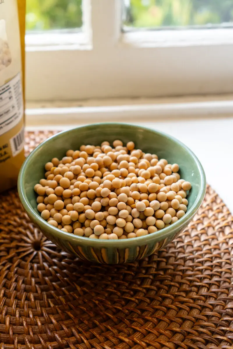 A bowl of dried soybeans. The soybeans sit in a green bowl on a woven mat. They sit on a window sill next to the bag. 