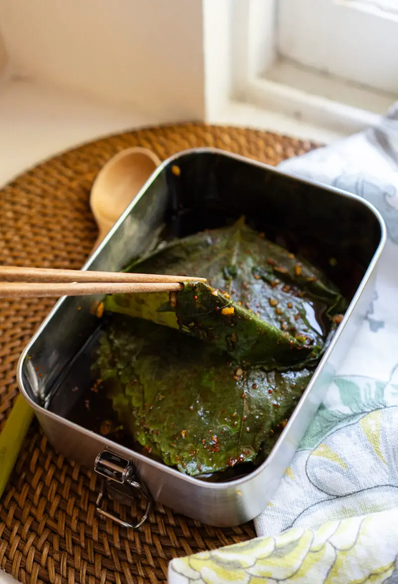 Korean pickled perilla leaves in a metal container. It sits on a table cloth next to spoon. I am lifting a perilla leaf with a wooden chopstick from the container. 