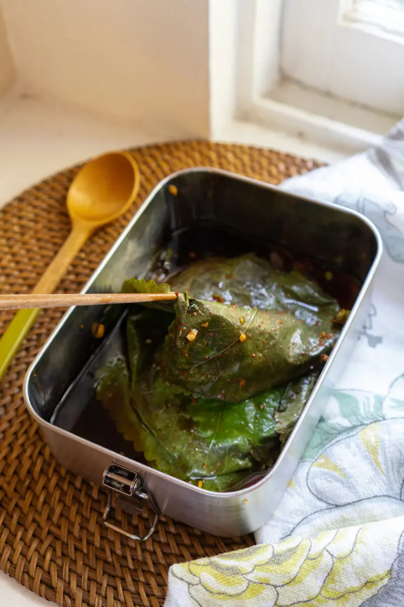 Kkaennip jangajji in a metal container. It sits on a table cloth next to spoon. I am lifting a perilla leaf with a wooden chopstick from the container. 