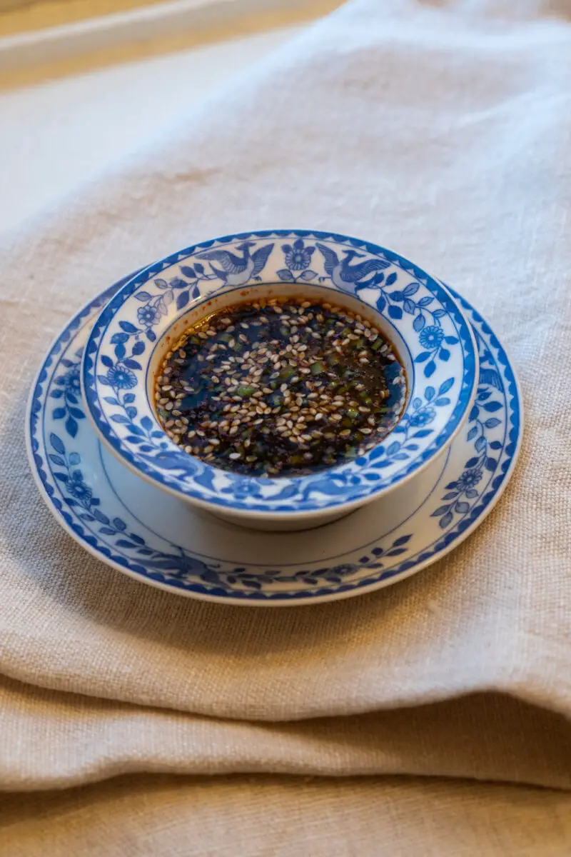A photo of Korean cho ganjang (jeon sauce) in a blue and white small bowl. The bowl sits on a matching plate. Both sit on a cream table cloth.