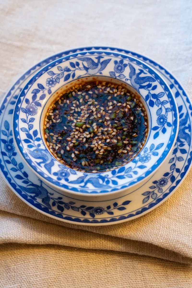 A side shot of Korean pancake sauce in a blue and white small bowl. The bowl sits on a matching plate. Both sit on a cream table cloth.