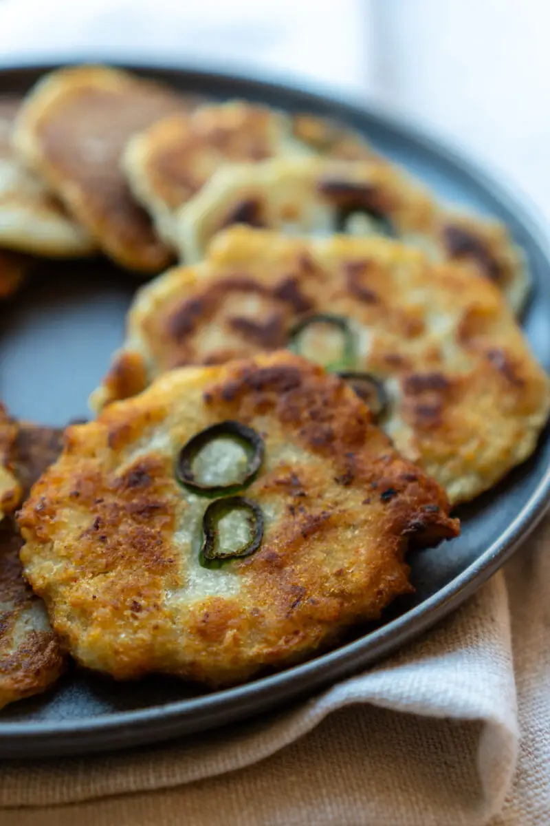 A closeup side shot of Korean gamja jeon on a plate. The round potato fritters have green hot peppers in the center. 