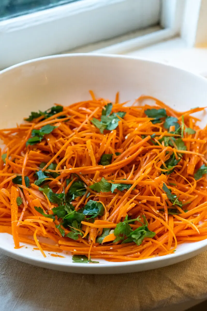 A closeup shot of Korean carrot salad in a white bowl. The salad has julienned carrots, spices, and cilantro. 
