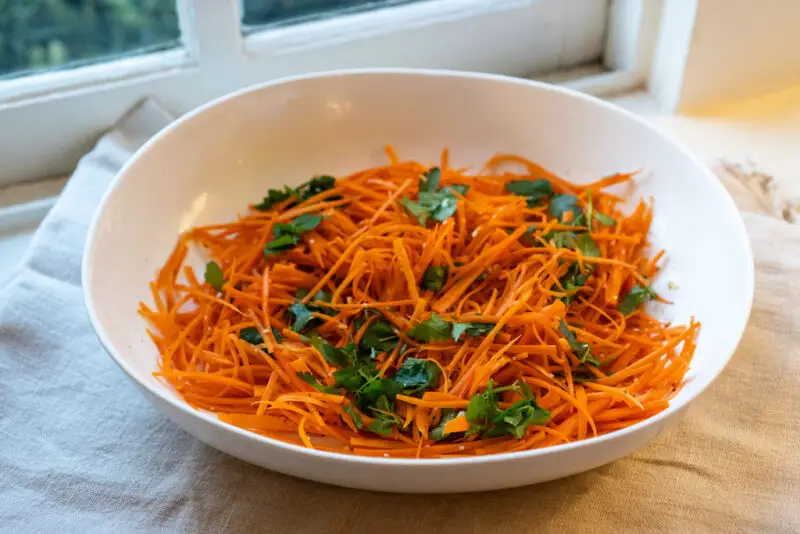 A shot of Korean carrot salad in a large white bowl on the window sill. The salad has julienned carrots, cilantro, spices, and more! 