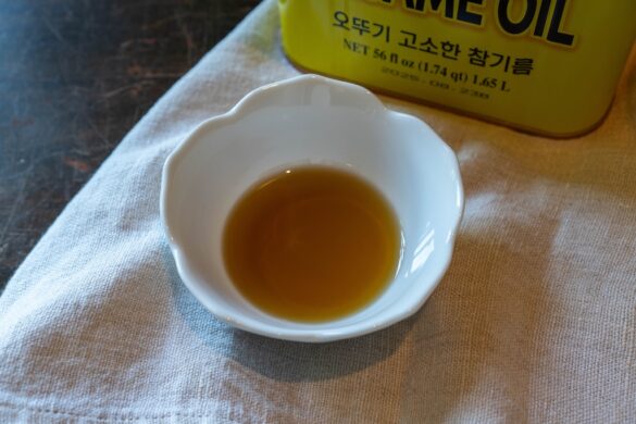 A closeup shot of a small white bowl of toasted sesame oil. A container of sesame oil sits in the background.