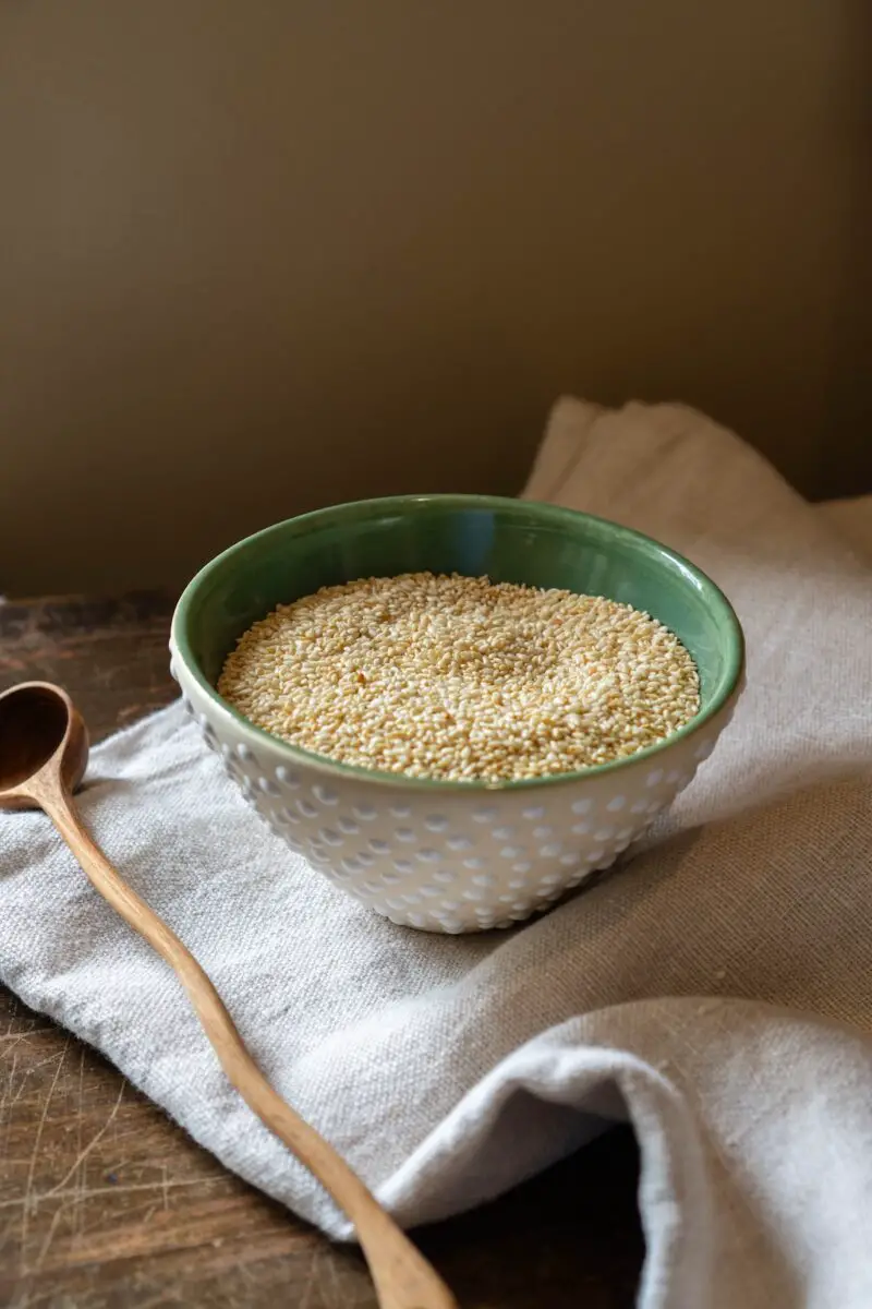 A side photo of sesame seeds sitting in a polkadot bowl with a green interior. The bowl sits on a table cloth next to a wooden spoon.