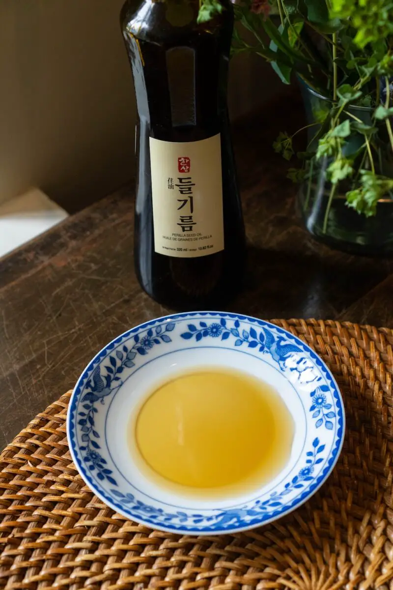 A photograph of perilla oil on a blue and white curved plate. The perilla oil bottle sits in the background. It sits on  a wicker place mat. 